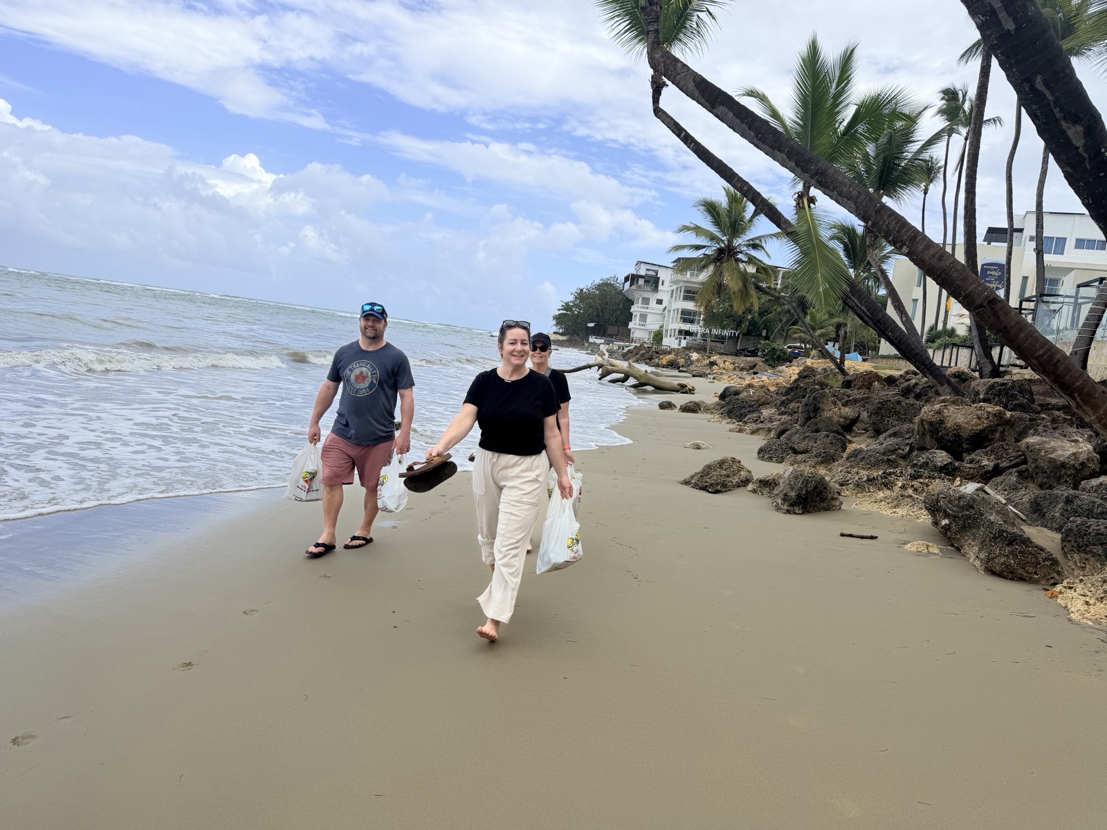 Three people walking barefoot along the sand carrying supermarket bags, palms leaning over the beach behind them