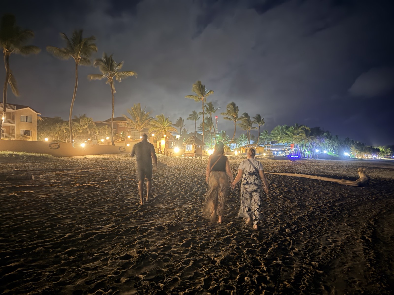 Three people walking home along the sand at night, with lit restaurants and palms ahead of them